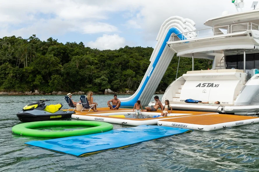 Produtos YouRoo em uso: plataformas flutuantes, escorregador inflável e piscina de mar ao lado de iate, com família aproveitando o lazer náutico